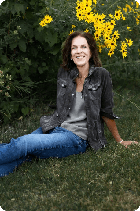 A woman is sitting in the grass in front of a bush with yellow flowers.