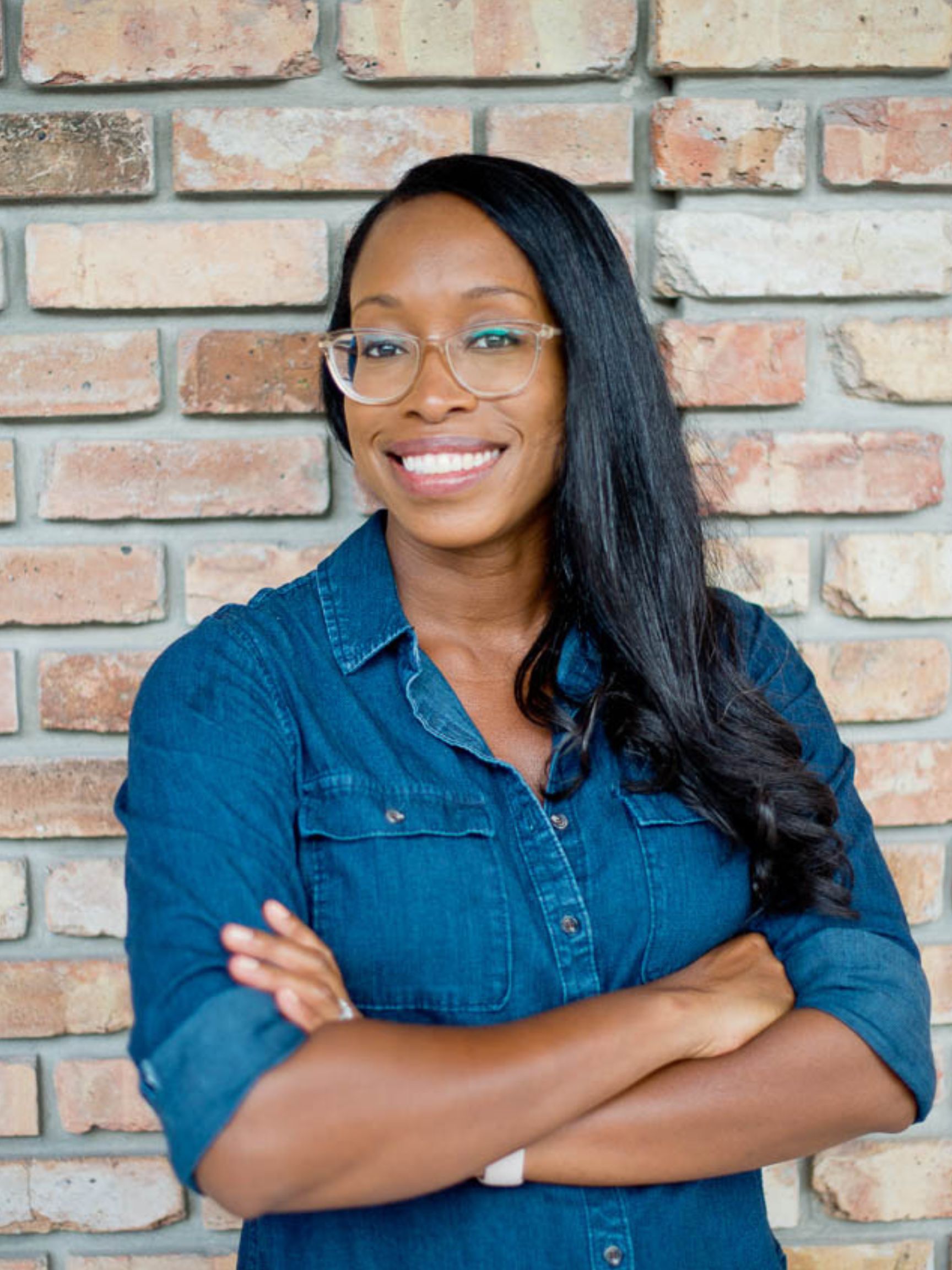 A woman wearing glasses and a blue shirt is standing in front of a brick wall.