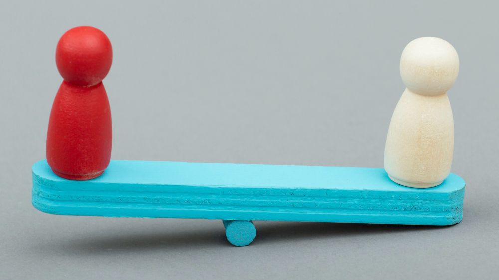 Two wooden figures are sitting on a blue seesaw.