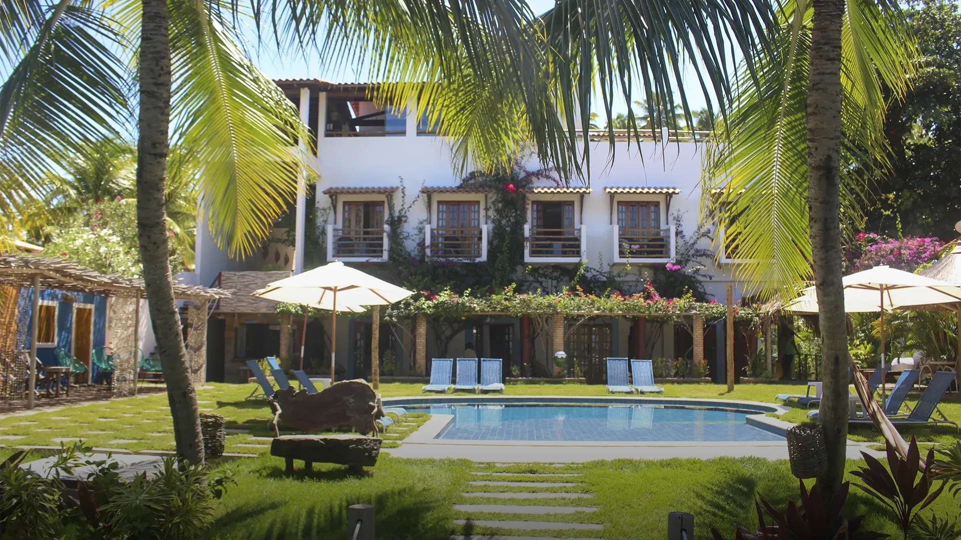 Resort tropical com piscina, prédio branco, palmeiras, céu azul.