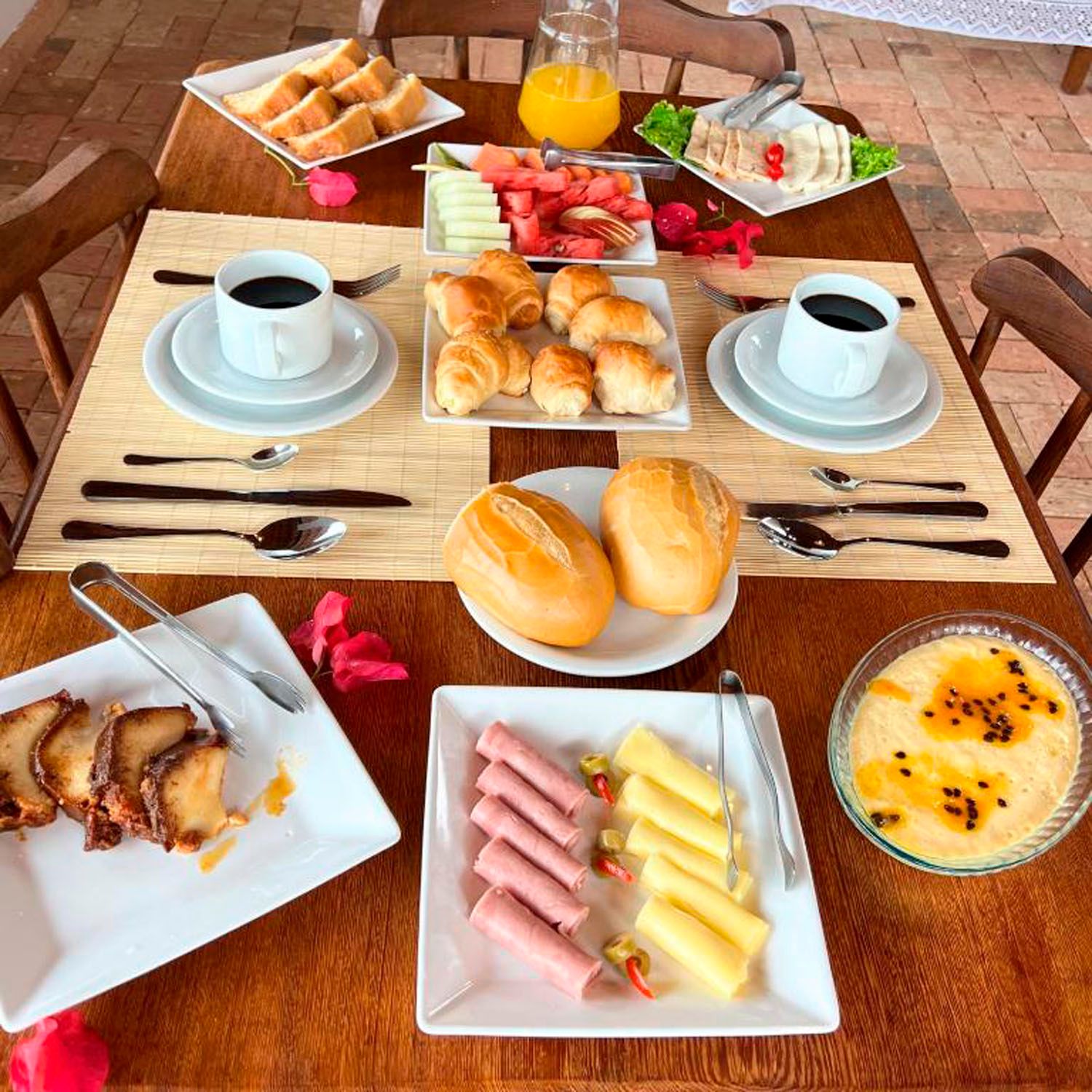 Café da manhã servido em uma mesa de madeira: café, frutas, doces, pão, presunto, queijo e sobremesa.