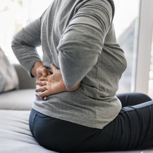 A woman is sitting on a couch holding her back in pain.