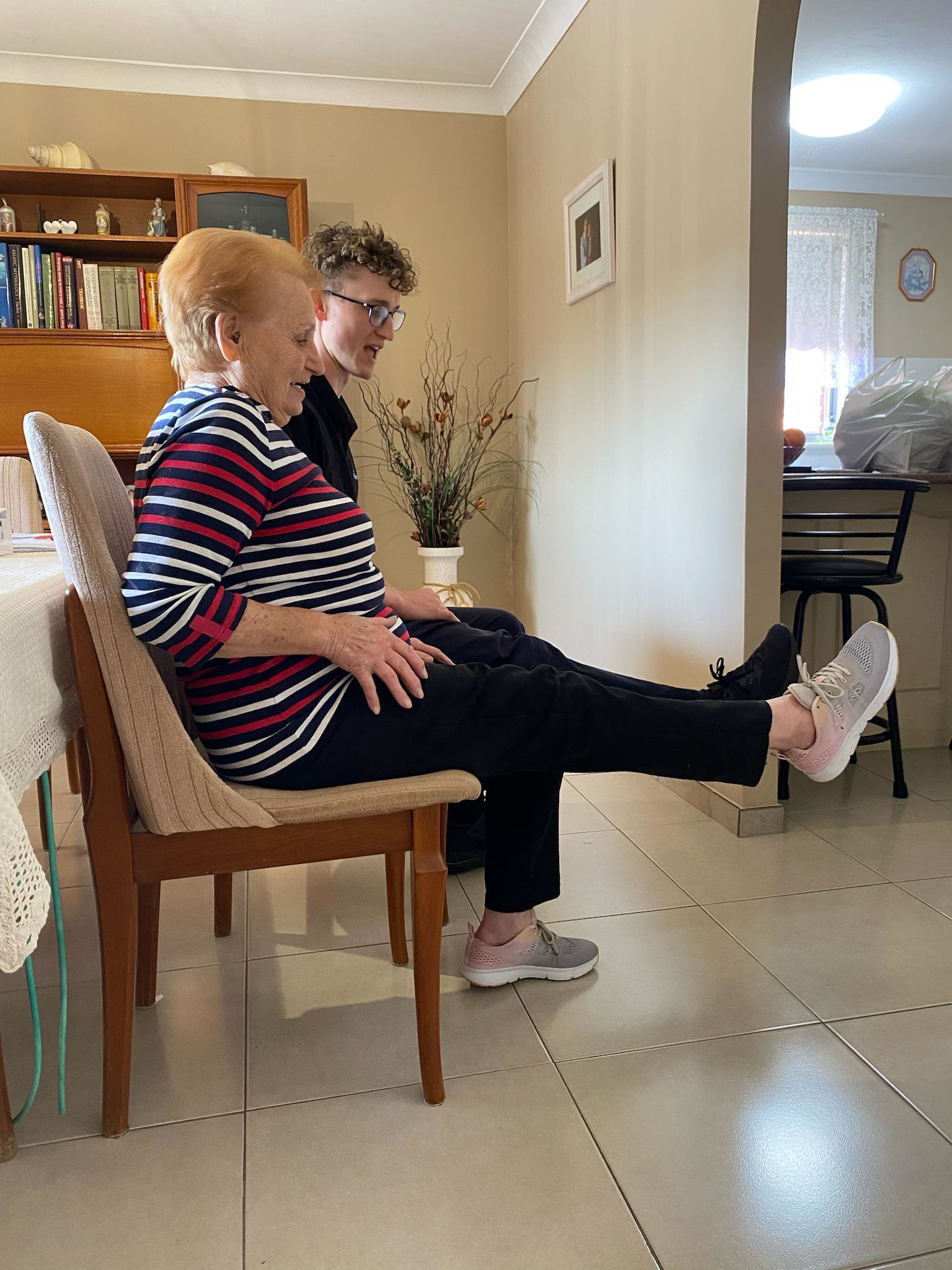 A boy is helping an elderly woman stretch her legs in a living room.