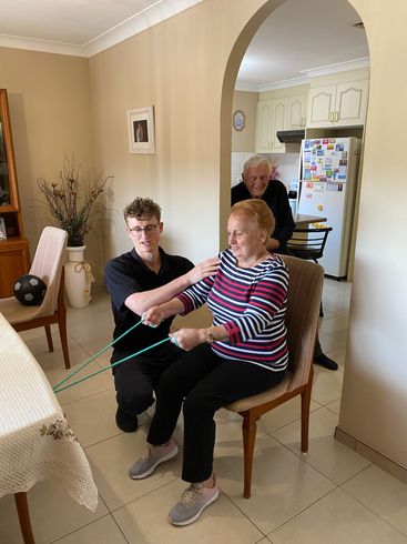 A man is helping an elderly woman with a resistance band in a living room.