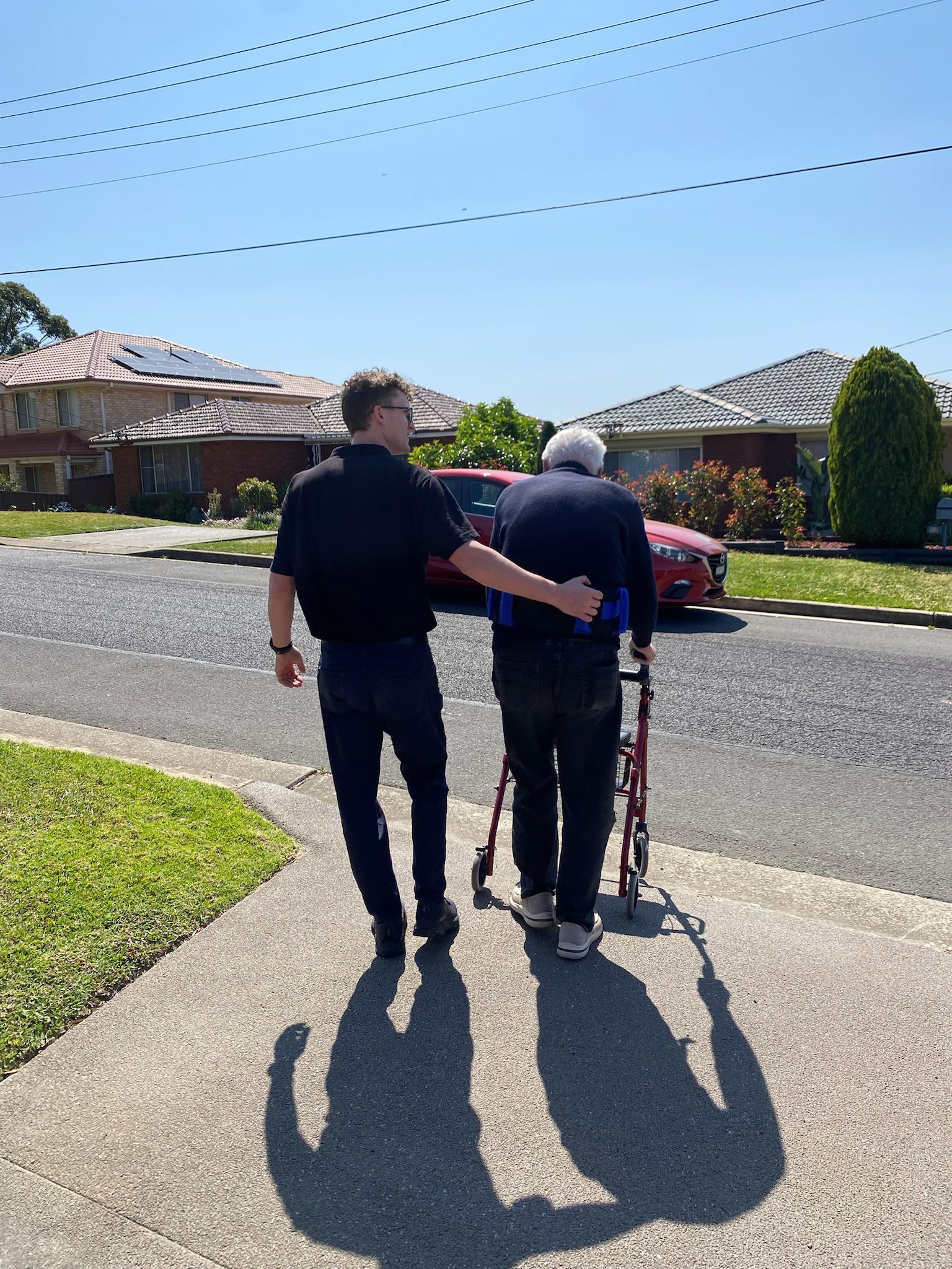 A man is helping an older man with a walker down a sidewalk.