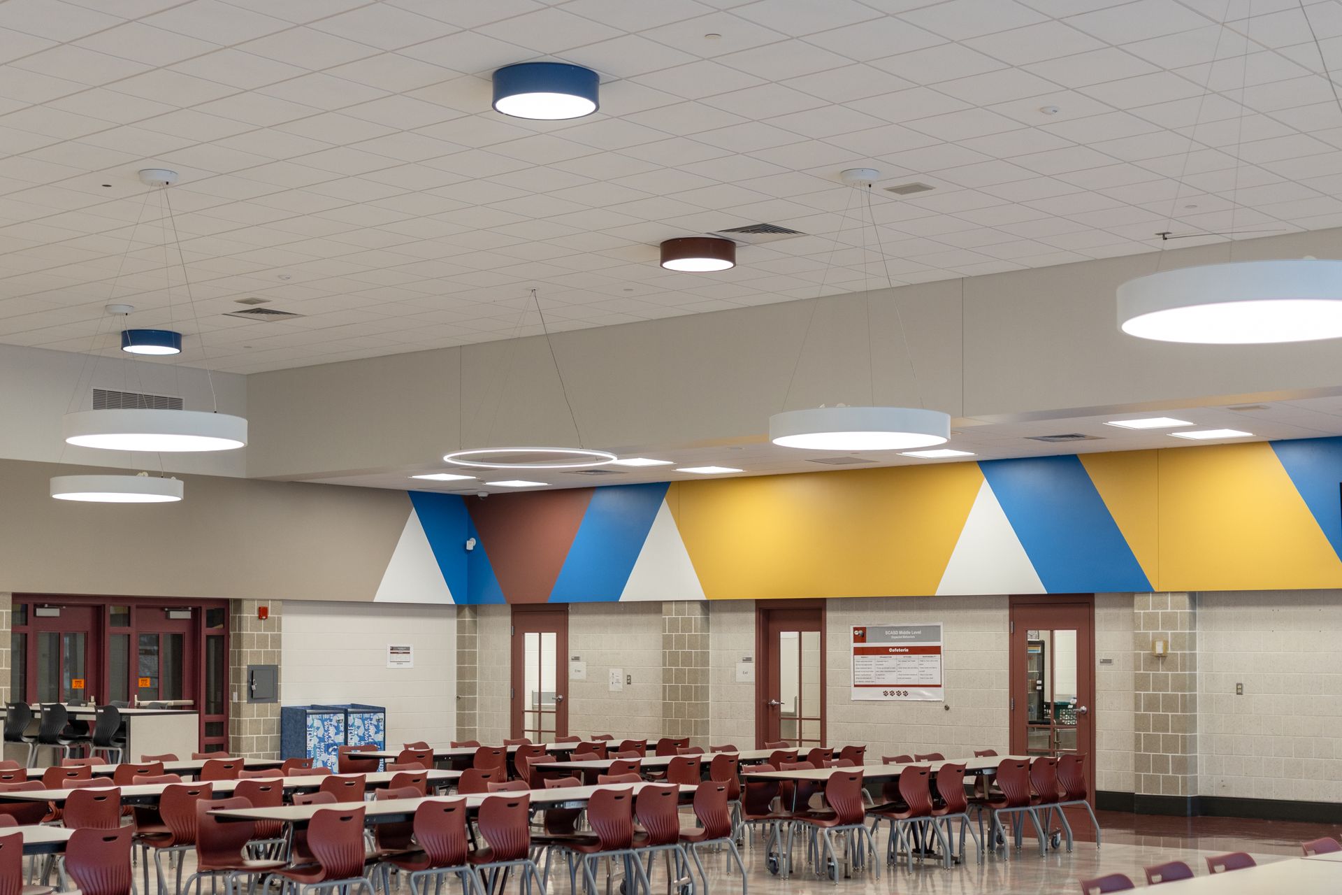 Mount Nittany Middle School Cafeteria with rows of red chairs, tables, and colorful wall panels under bright ceiling lights