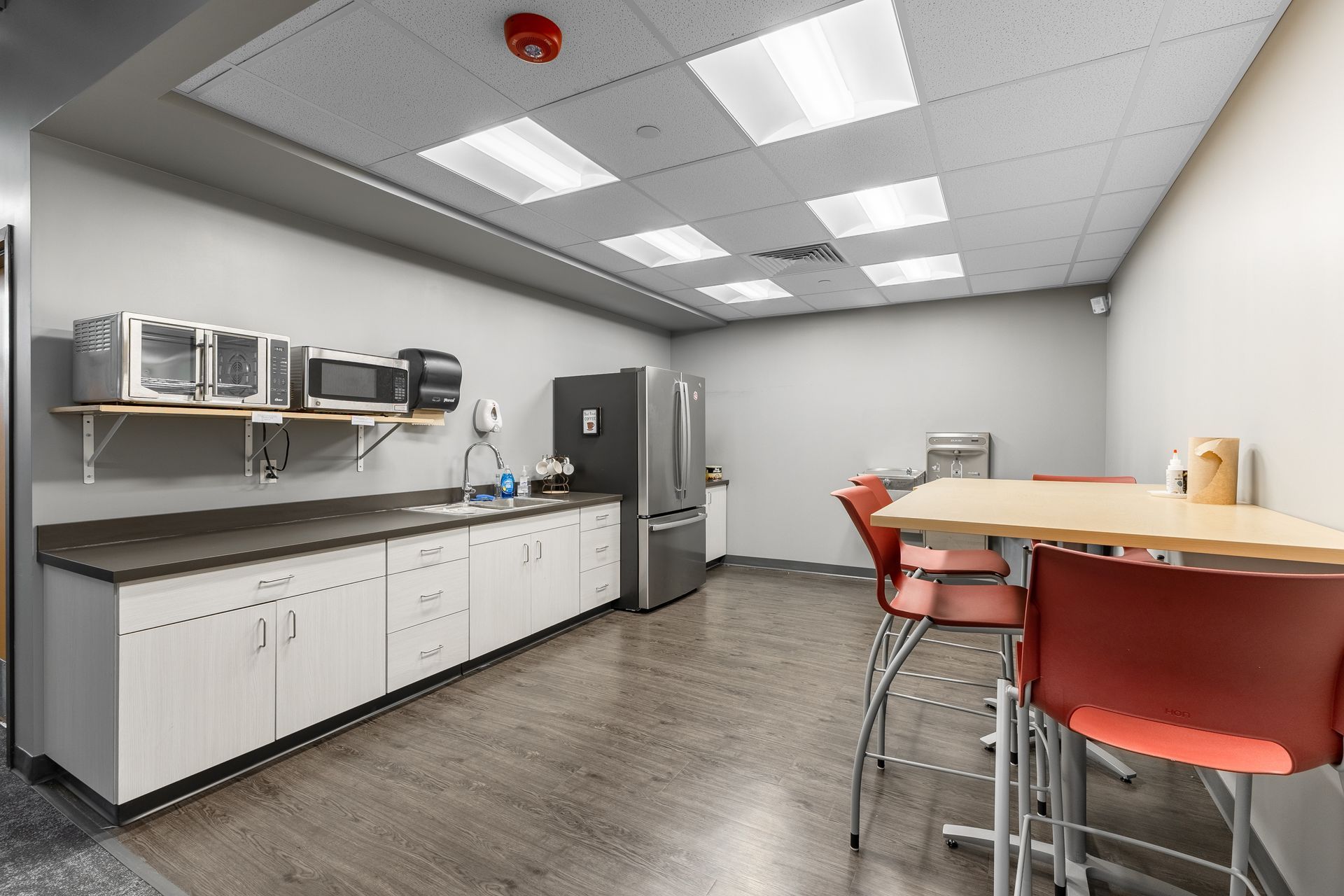 State College Area School District North Building's break room with cabinets, microwave, refrigerator, table, and red bar stools under bright ceiling lights