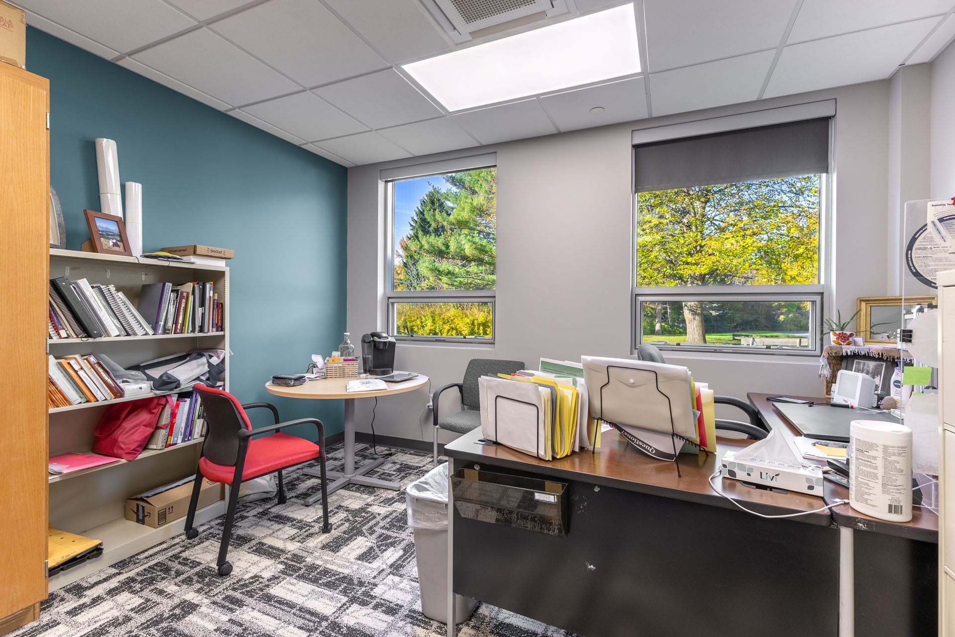 State College Area School District Panorama Village building office with desks, shelves, red chair, and sunlight through windows with yellow trees outside