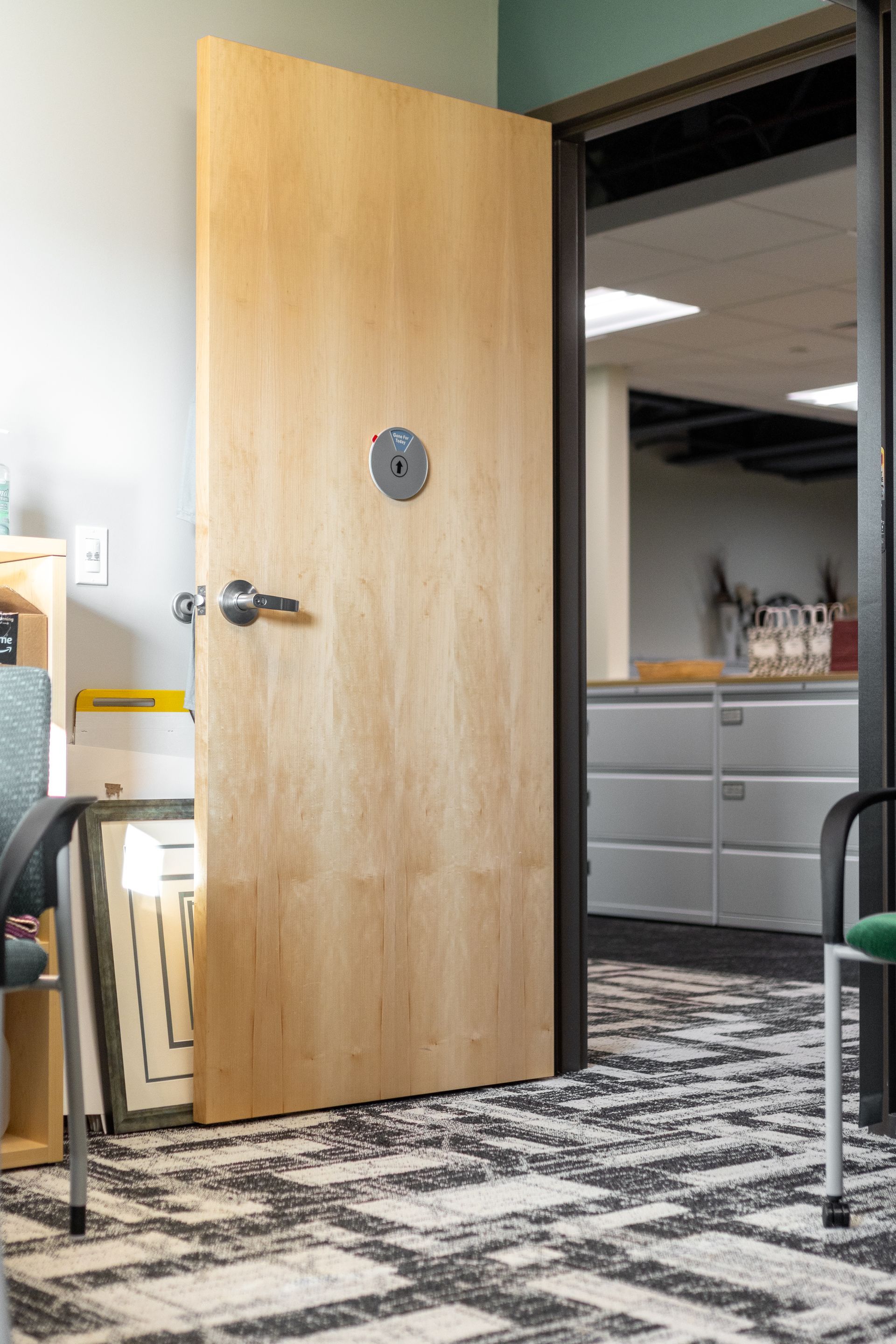 State College Area School District Panorama Village building's open wooden office door with round window, revealing a cubicle workspace with gray cabinets and black-and-white carpet.