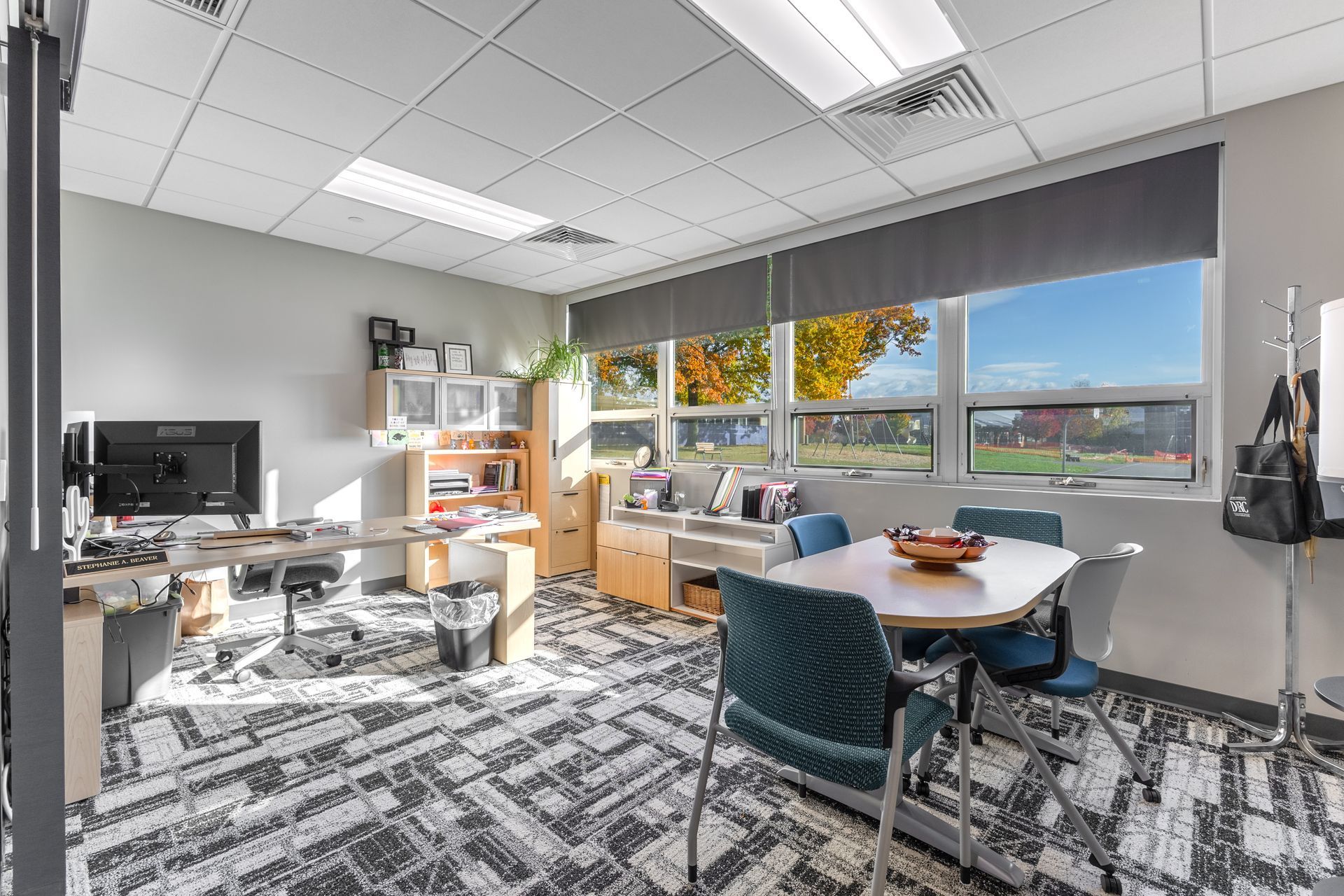 State College Area School District Panorama Village Building Office with desks, chairs, computers, and large windows overlooking autumn trees and a parking lot
