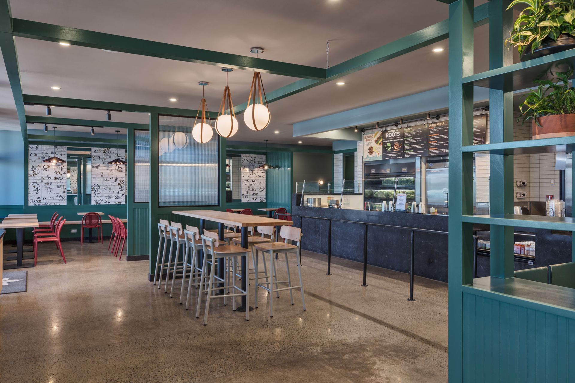 Roots Natural Kitchen interior with teal framework, light wood tables, metal stools, pendant lights, and concrete floors.