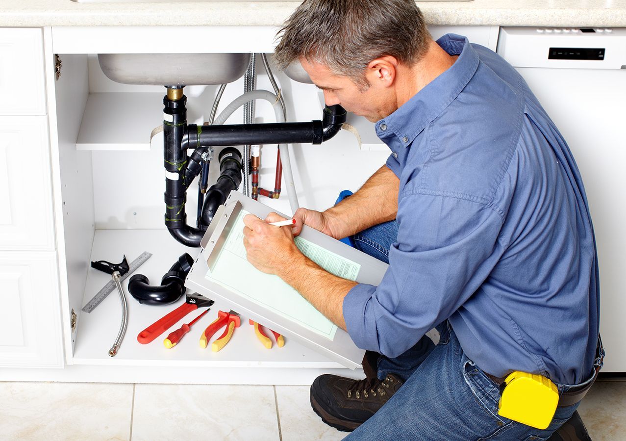 A plumber is fixing a sink in a kitchen and writing on a clipboard.