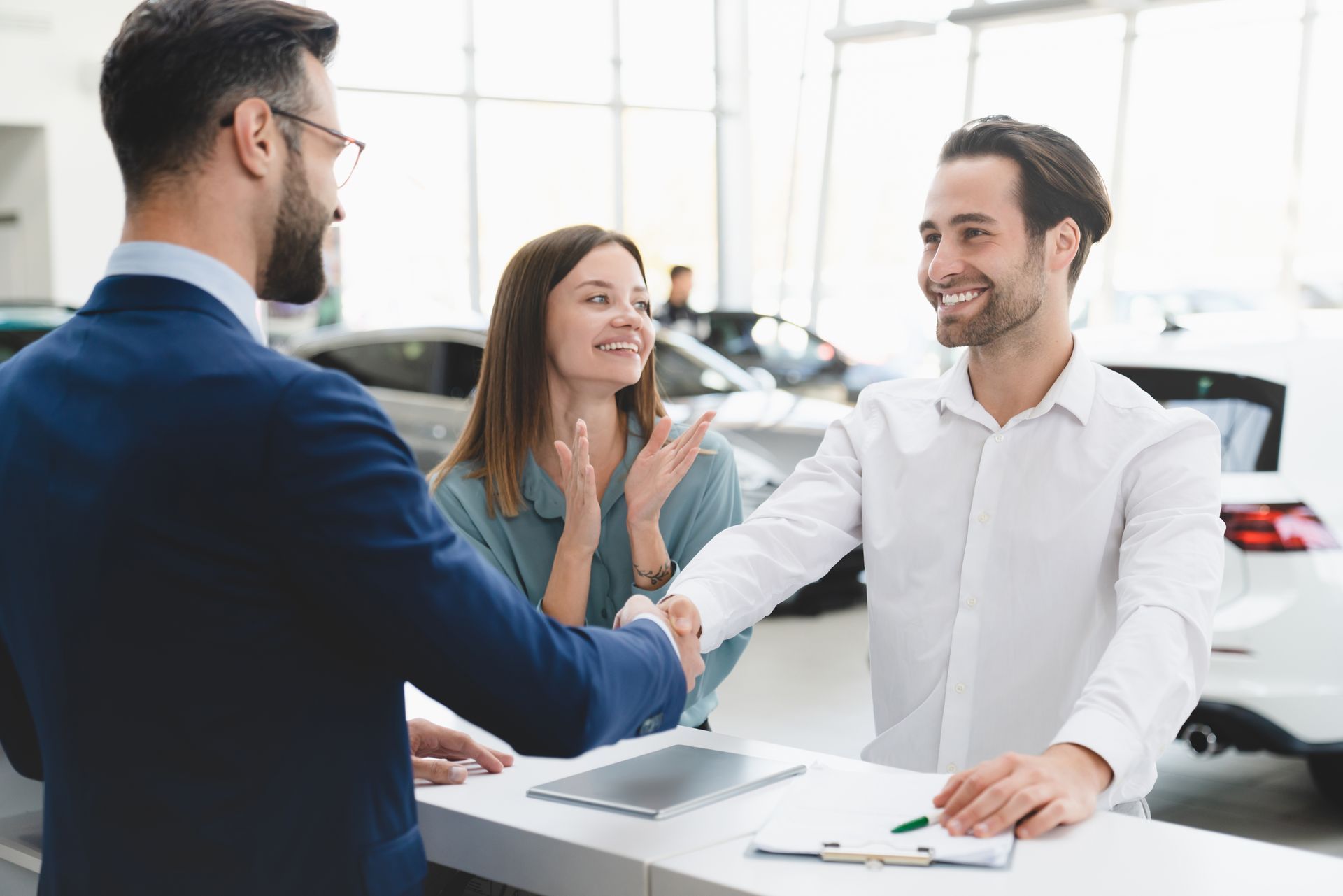 Car salesperson shaking hands with a customer; happy couple watches at dealership.