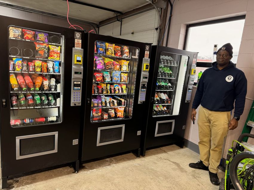 Man standing beside a lit vending machine stocked with drinks and snacks indoors.