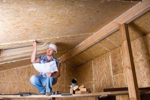 Male builder reads plans while sitting on top of upper crawlspace of an unfinished home.