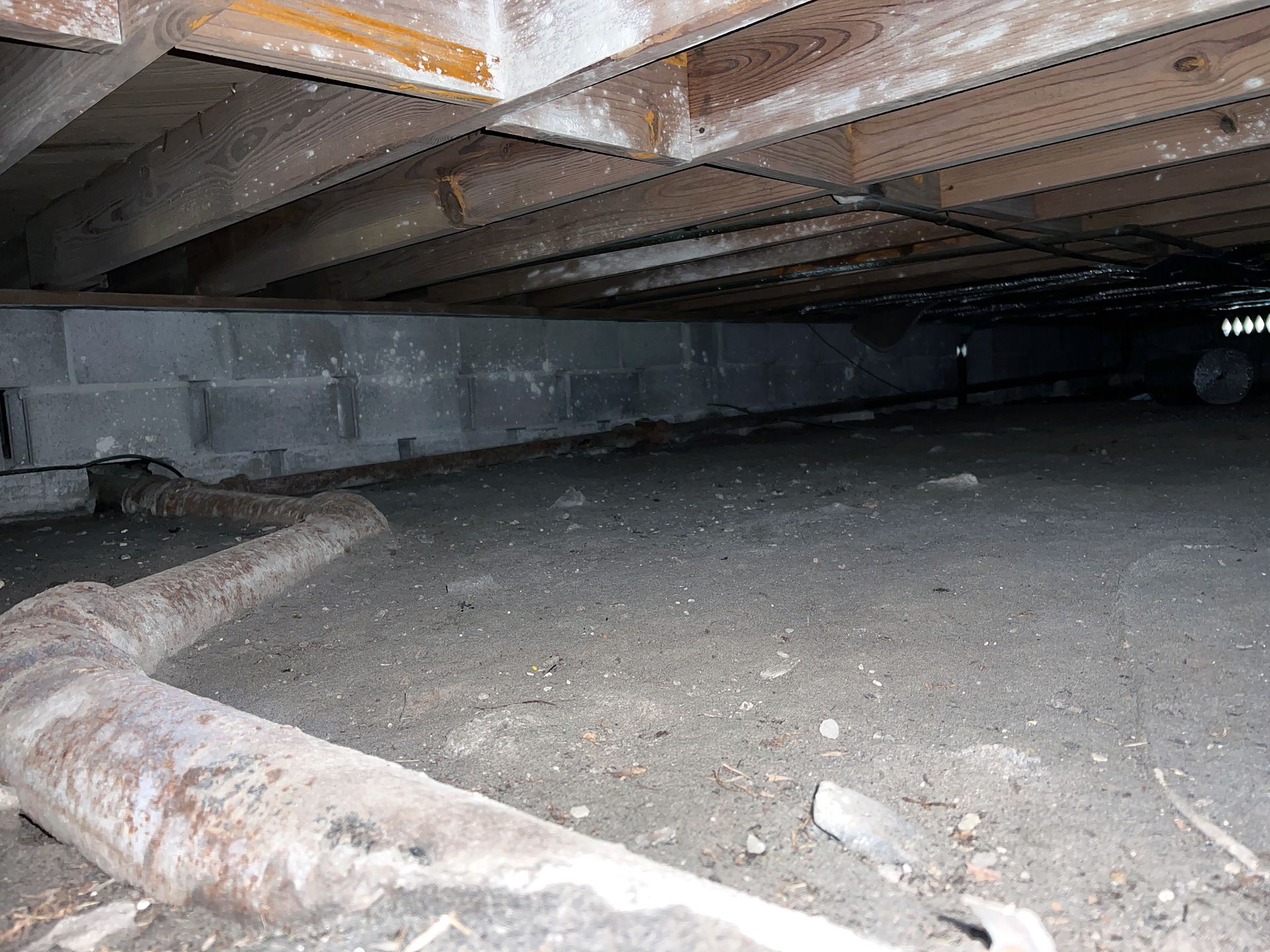 A low crawl space with exposed pipes, concrete walls, and wooden floor joists overhead.