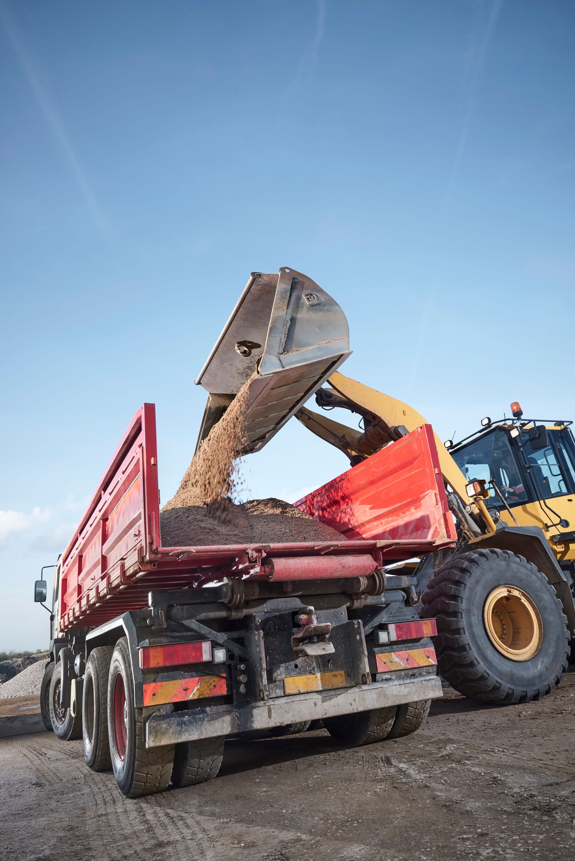 Yellow excavator dumping gravel into a red dump truck, outdoors.