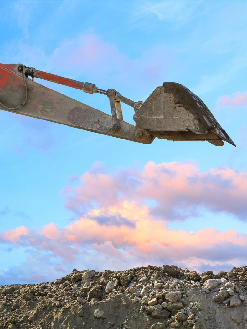 An excavator's bucket scoops earth against a backdrop of a cloudy, blue sky.