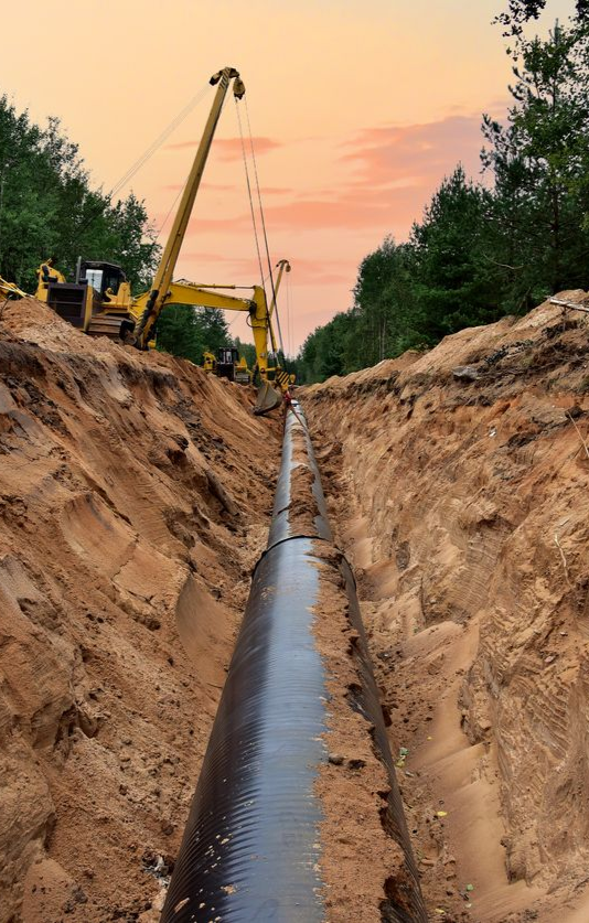 Pipeline being installed in a trench by yellow construction equipment, in a forest, at sunset.