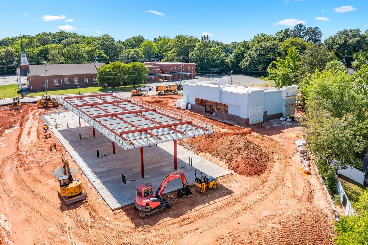 Construction site with steel framework and heavy machinery, with buildings and trees in the background.