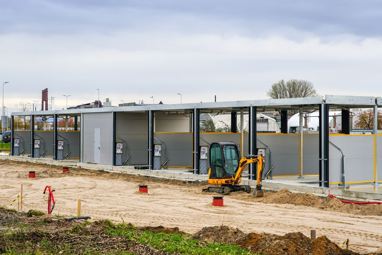 Construction site with car wash bays, yellow excavator, dirt ground, overcast sky.