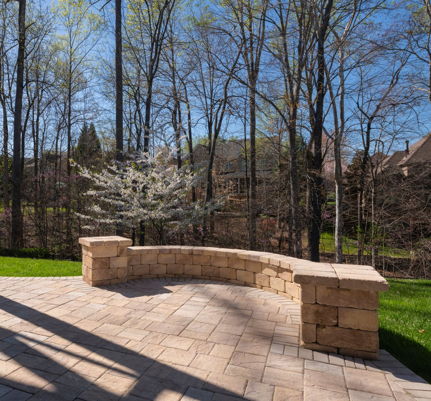 A curved stone retaining wall sits on a light-colored paver patio overlooking a wooded area with a blooming white tree.
