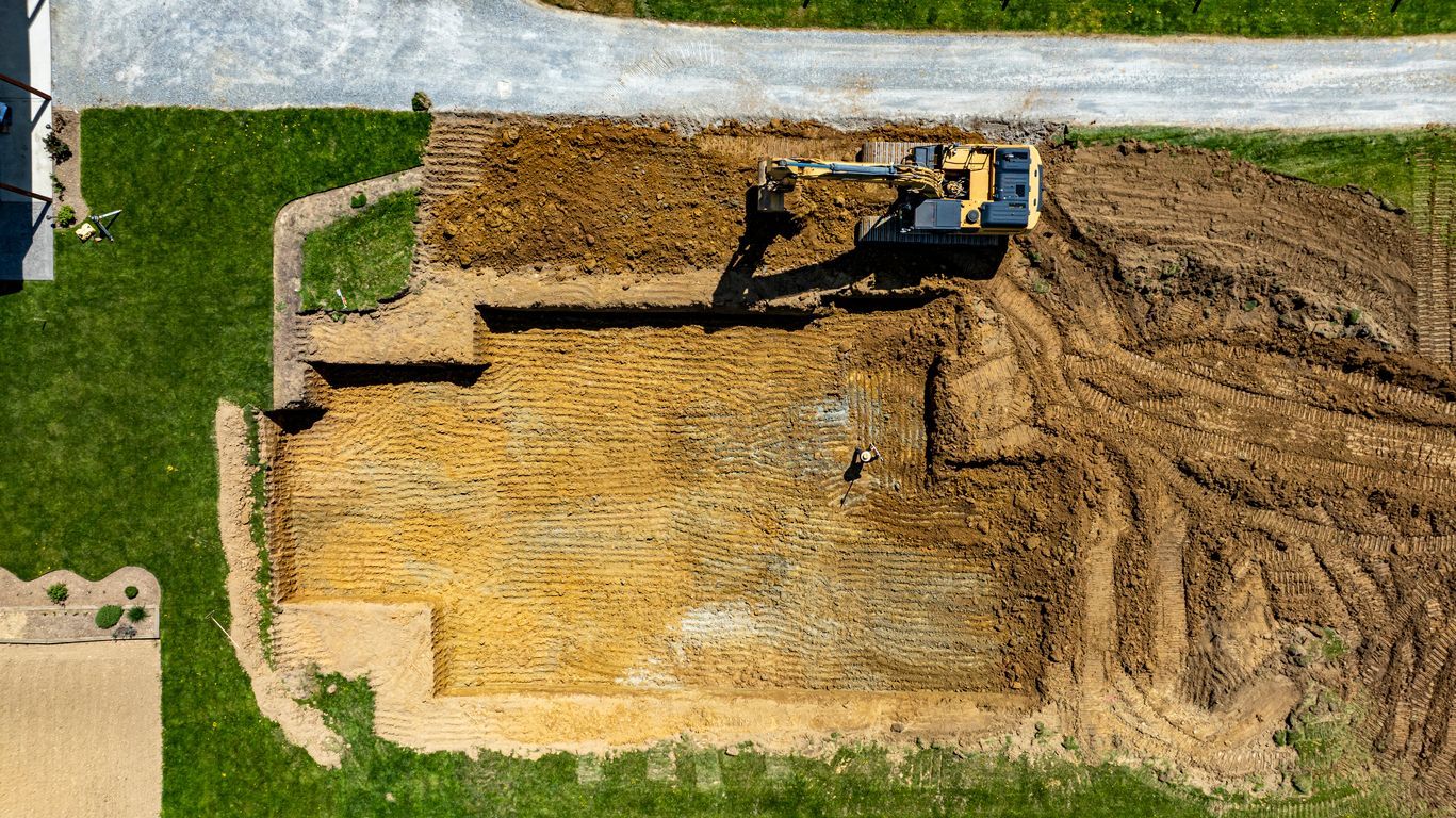 Overhead view of a construction site with an excavator digging into the earth; nearby a patch of grass and dirt road.