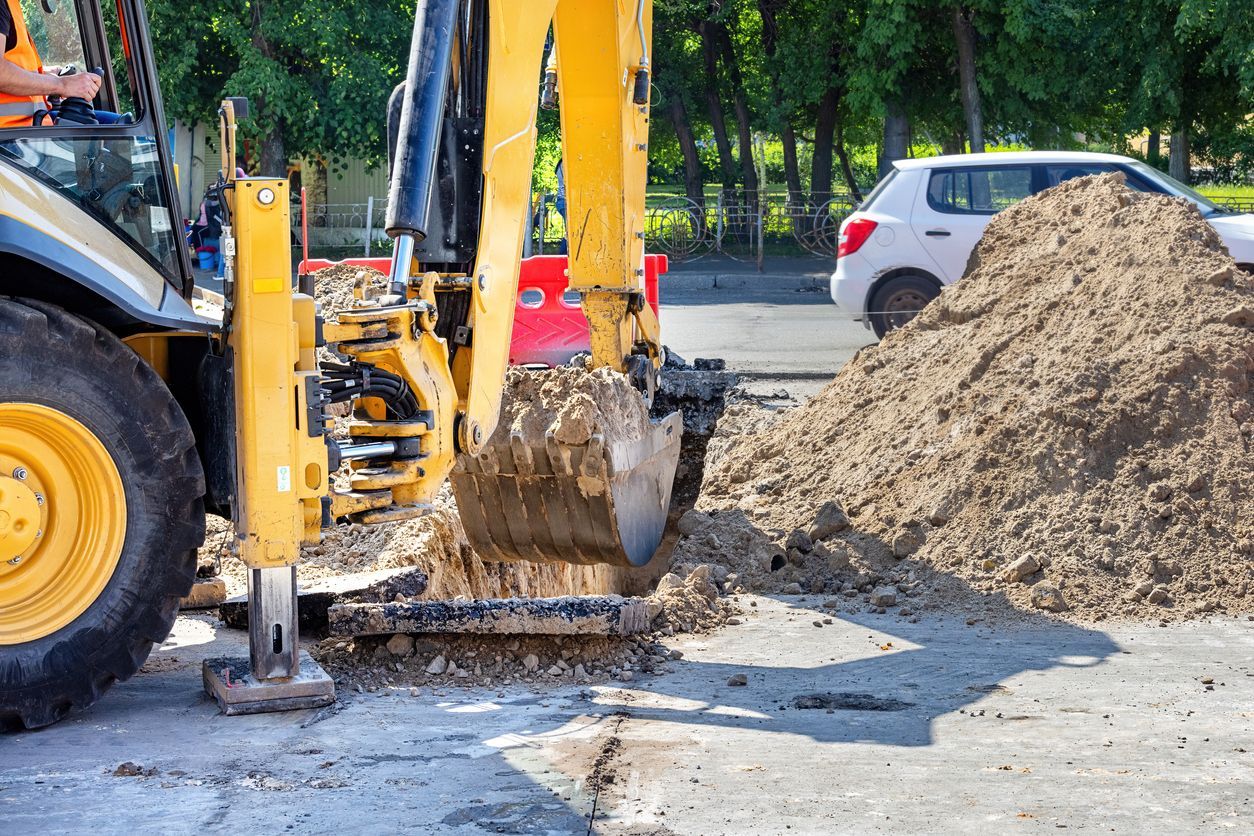 Yellow backhoe digging on a road with a dirt pile and a white car in the background.