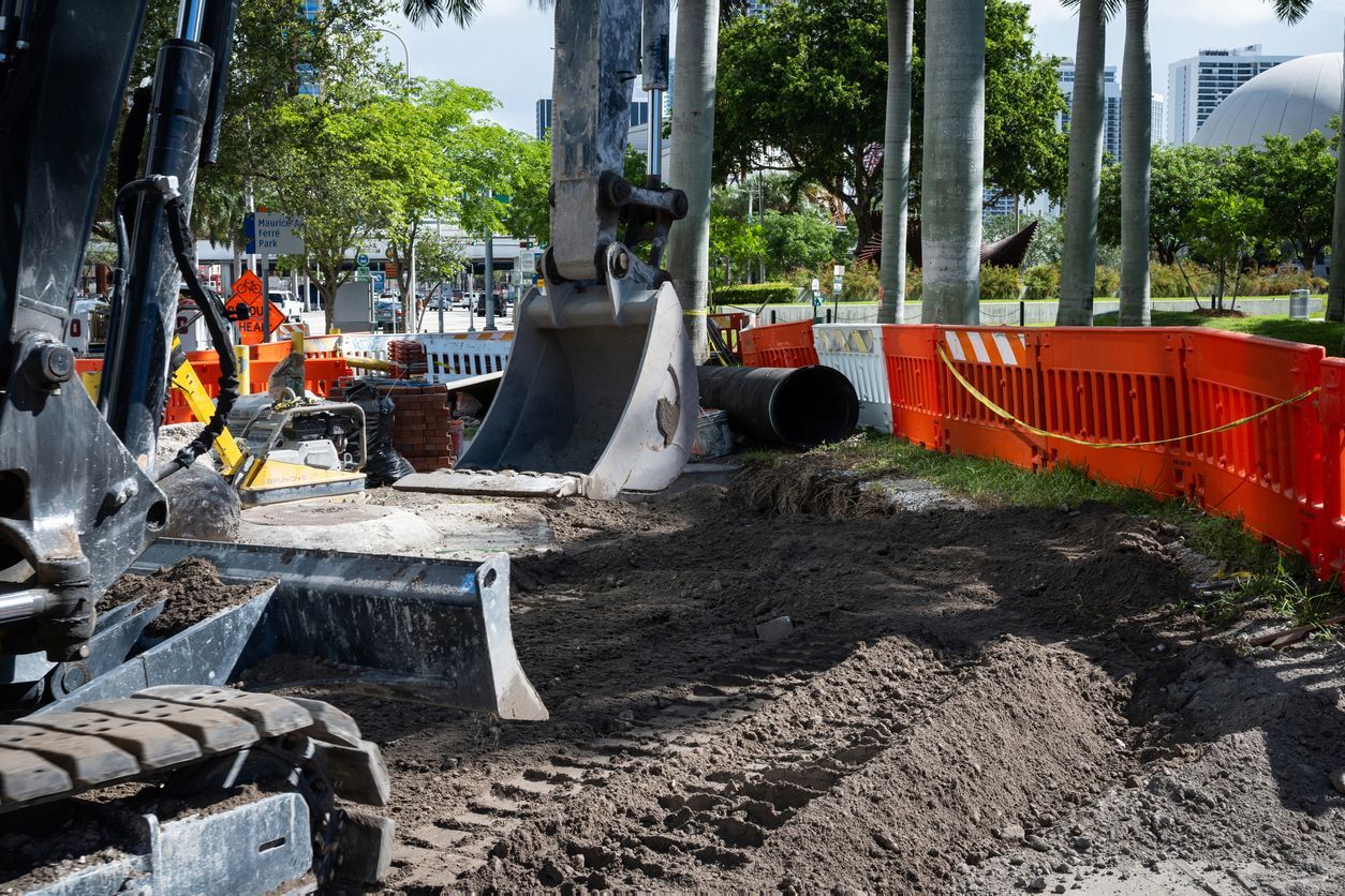 Excavator digging in dirt at a construction site, orange barriers, trees in background.