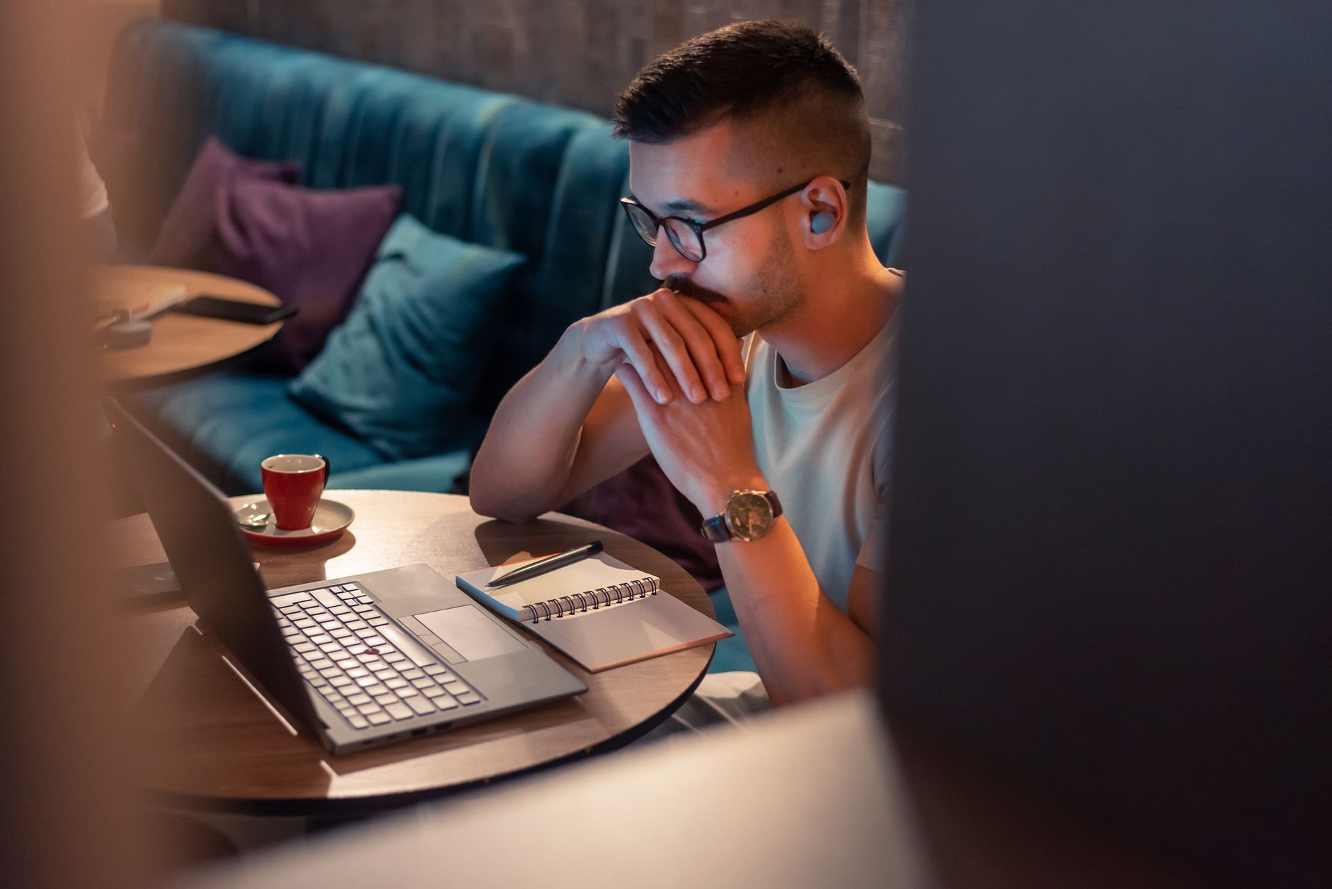 Man at a table with a laptop, notebook, and coffee, looking at the screen with a focused expression.