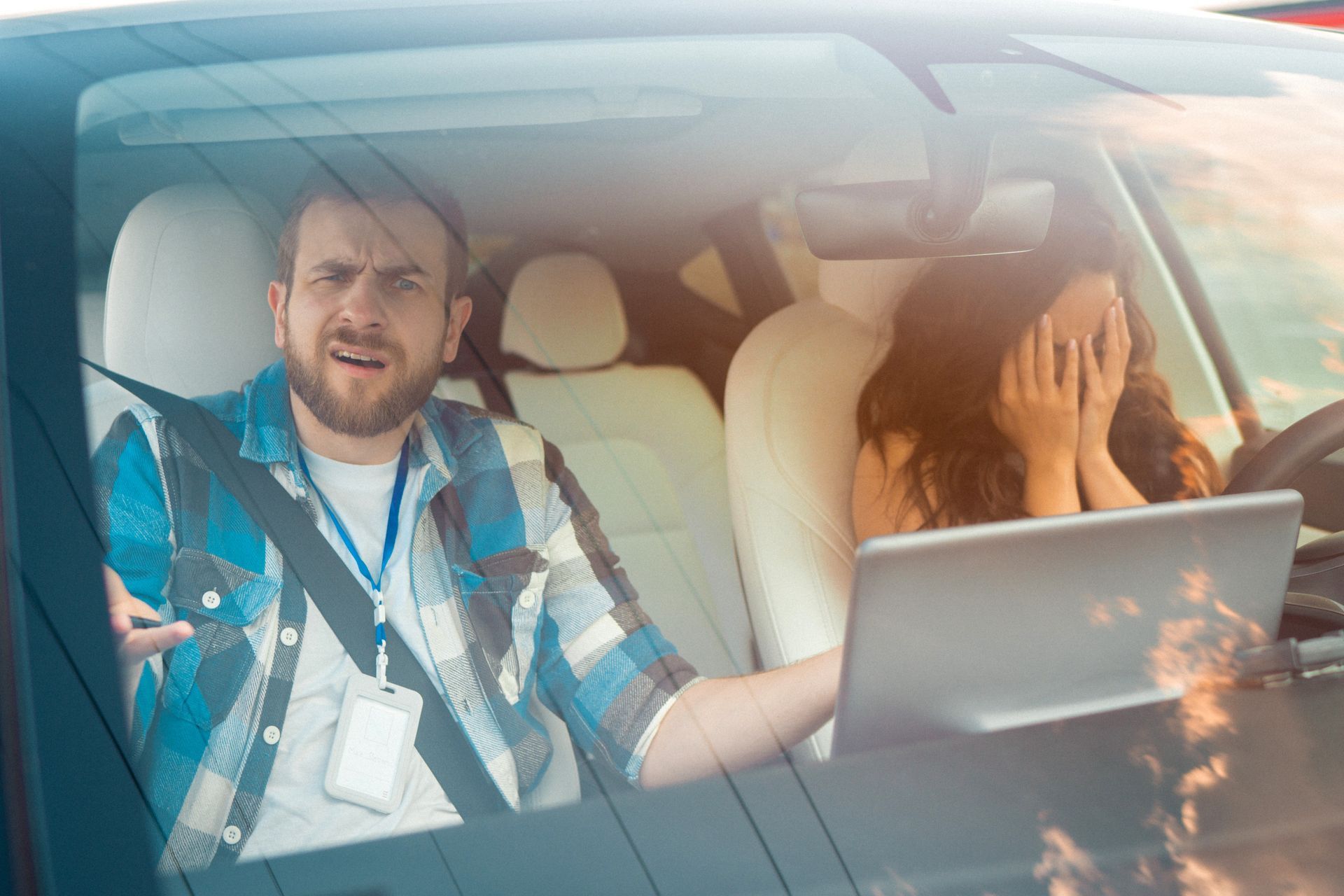Man in plaid shirt gestures angrily at woman covering face, laptop on steering wheel, in car.