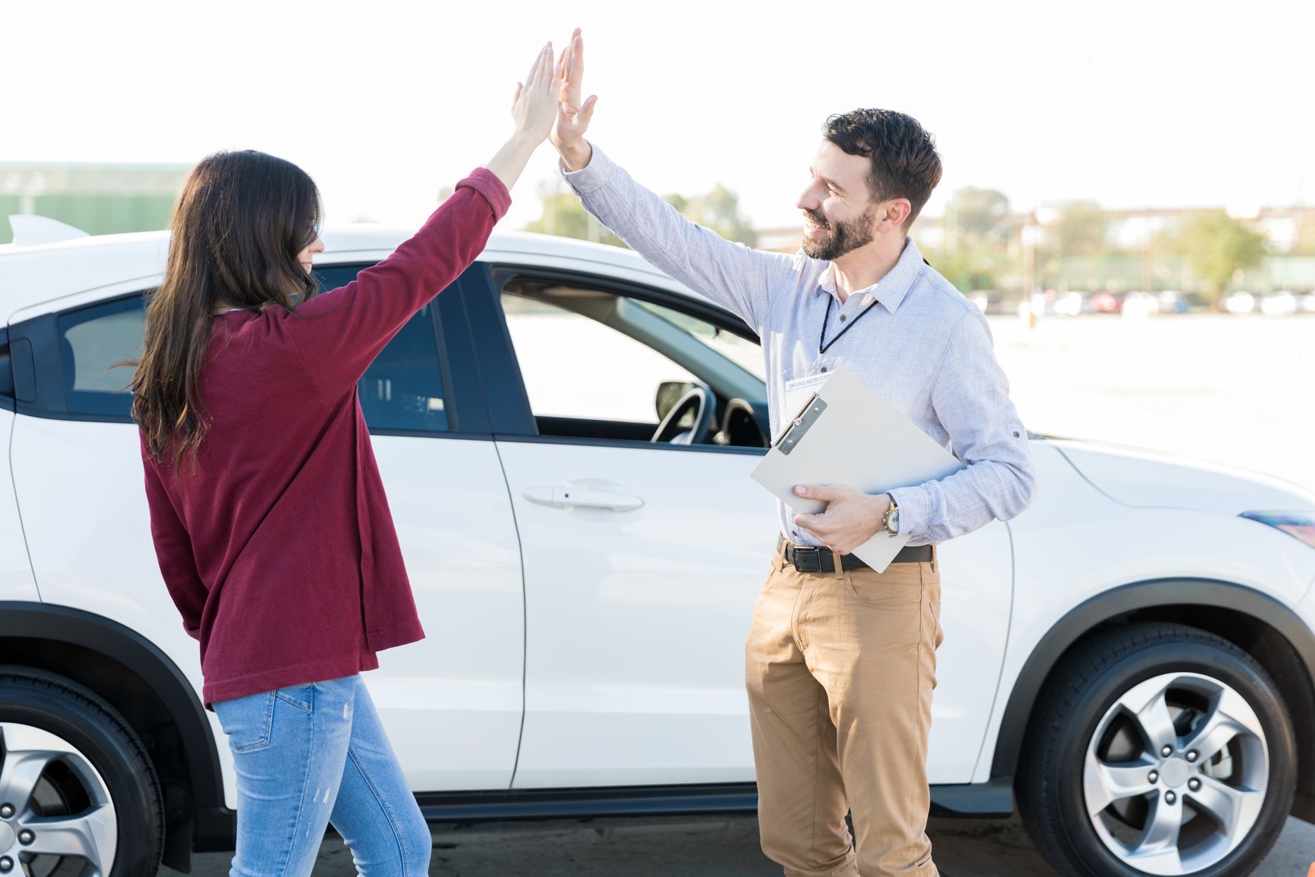 Woman and instructor high-fiving by white car; instructor holds a clipboard. Woman and instructor high-fiving by white car; instructor holds a clipboard.