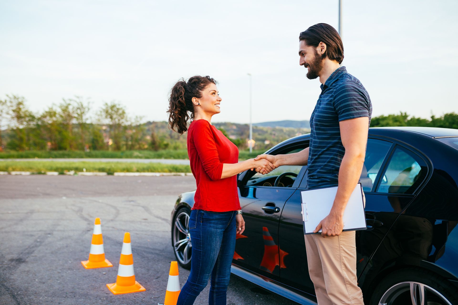Woman in red shirt shakes hands with man holding clipboard next to black car and traffic cones.