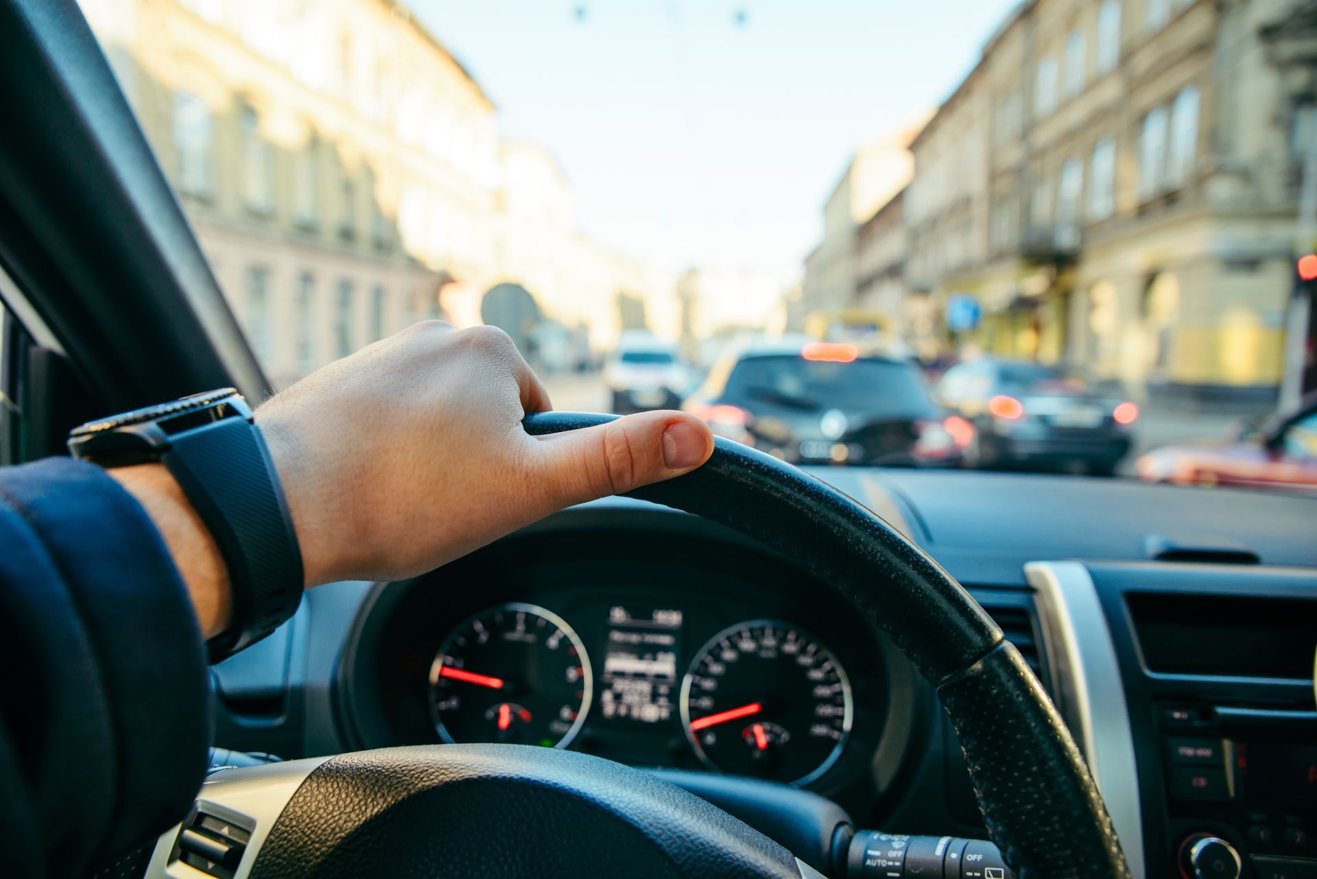 Hand gripping steering wheel driving through a city street with cars and buildings. Hand gripping steering wheel driving through a city street with cars and buildings.