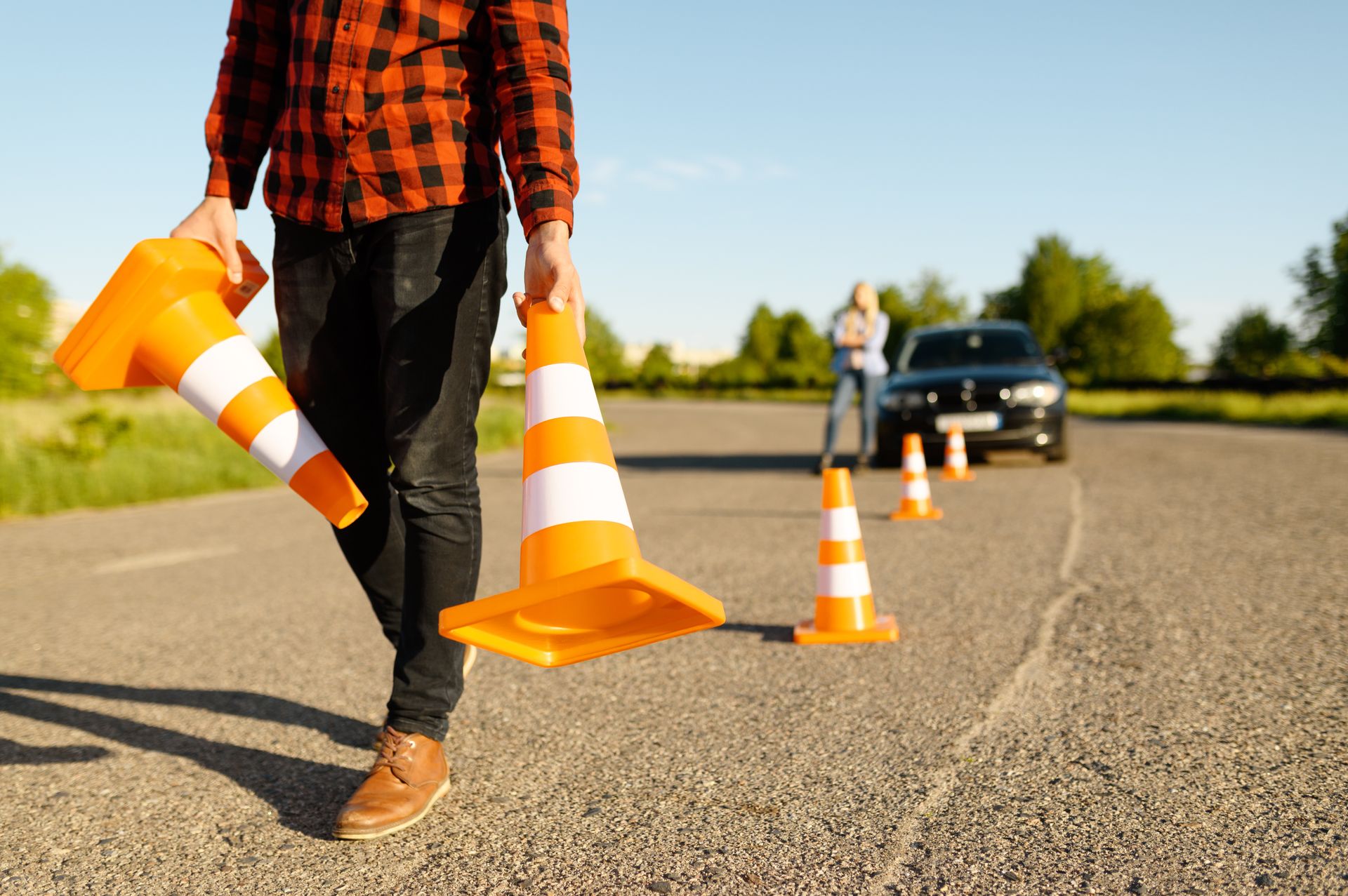Person carrying orange traffic cones, setting up driving course. A car is in the background.
