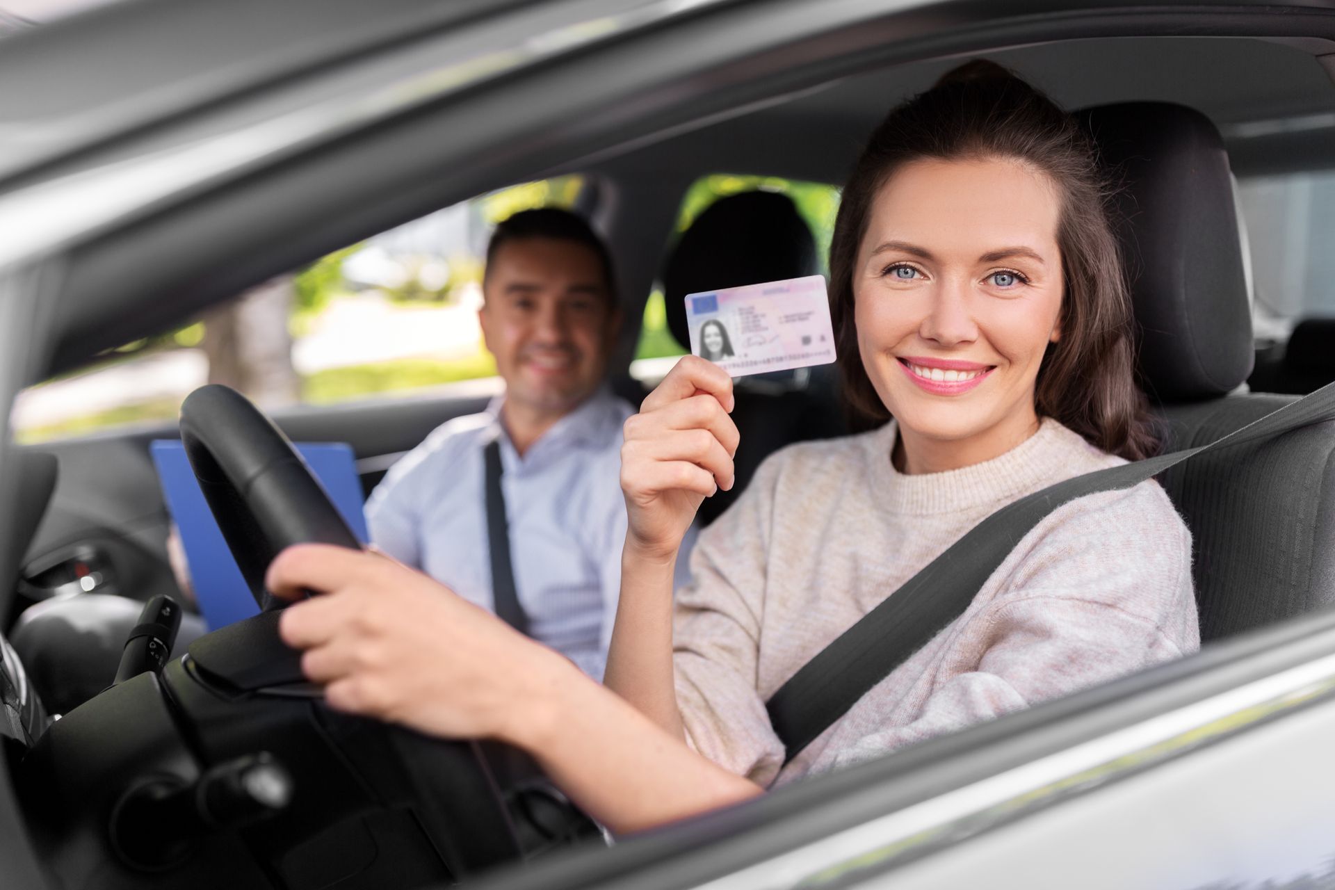 Woman in car holding driver's license, smiling. Instructor in passenger seat smiles.