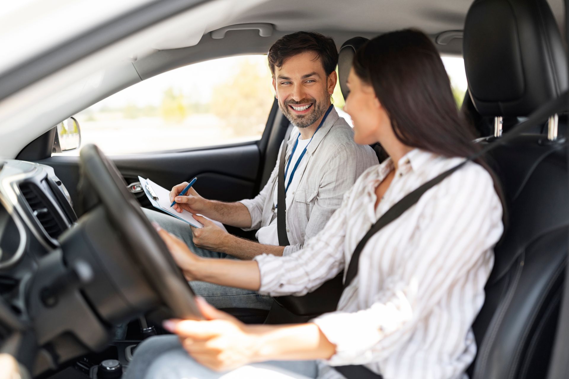 Driving instructor smiling at a driver during a lesson, both in a car, instructor holding a clipboard. Driving instructor smiling at a driver during a lesson, both in a car, instructor holding a clipboard.