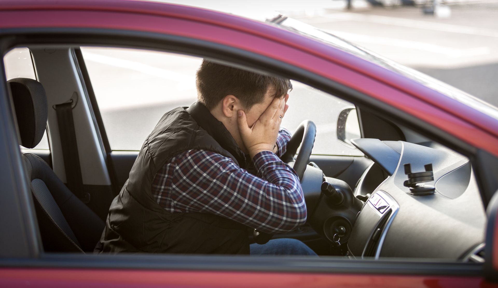 Man in a red car, covering his face with hands, looking stressed.