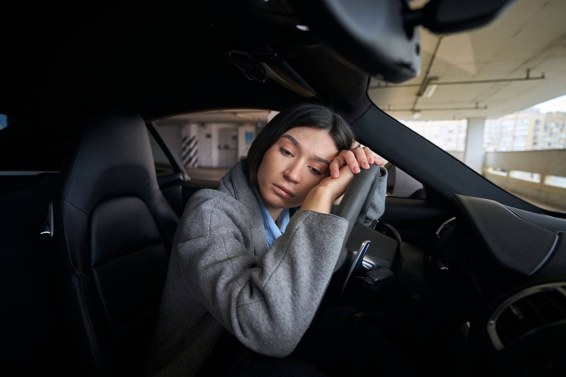 Woman resting her head on a steering wheel inside a dark car, appearing tired in a parking garage. Woman resting her head on a steering wheel inside a dark car, appearing tired in a parking garage.