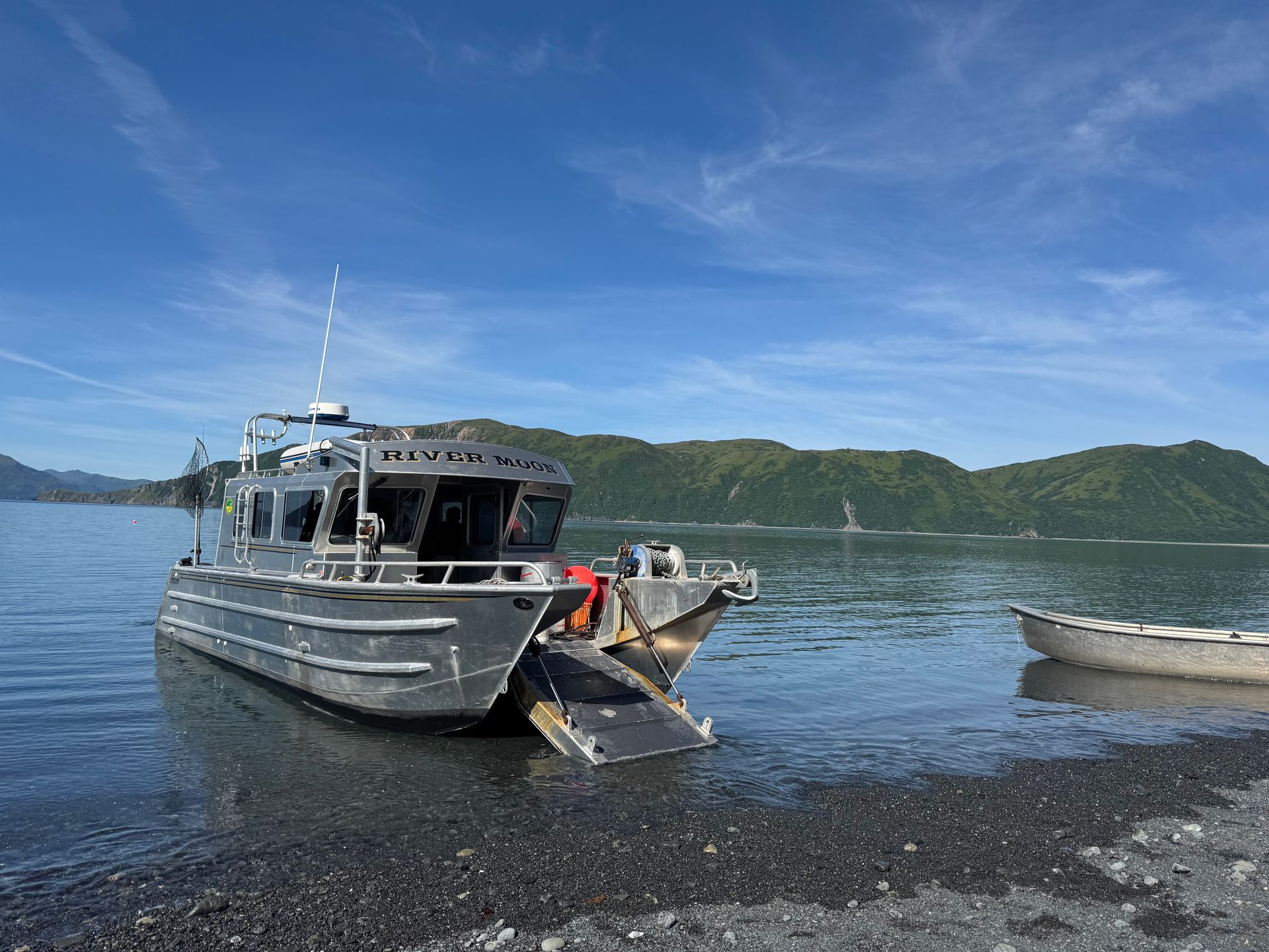 A silver boat with a ramp on a rocky shore, under a blue sky. A small boat floats nearby.