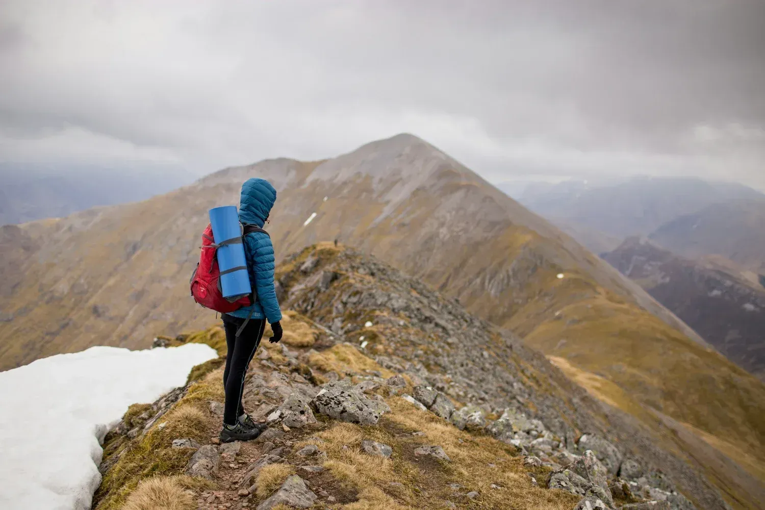Person with backpack stands on a mountain ridge, looking at the cloudy landscape.