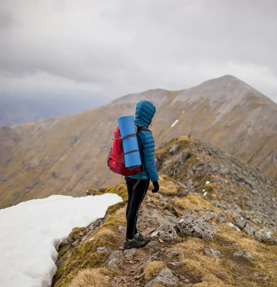 Hiker stands on a mountain ridge, gazing at a peak. Red backpack, blue jacket, cloudy sky.