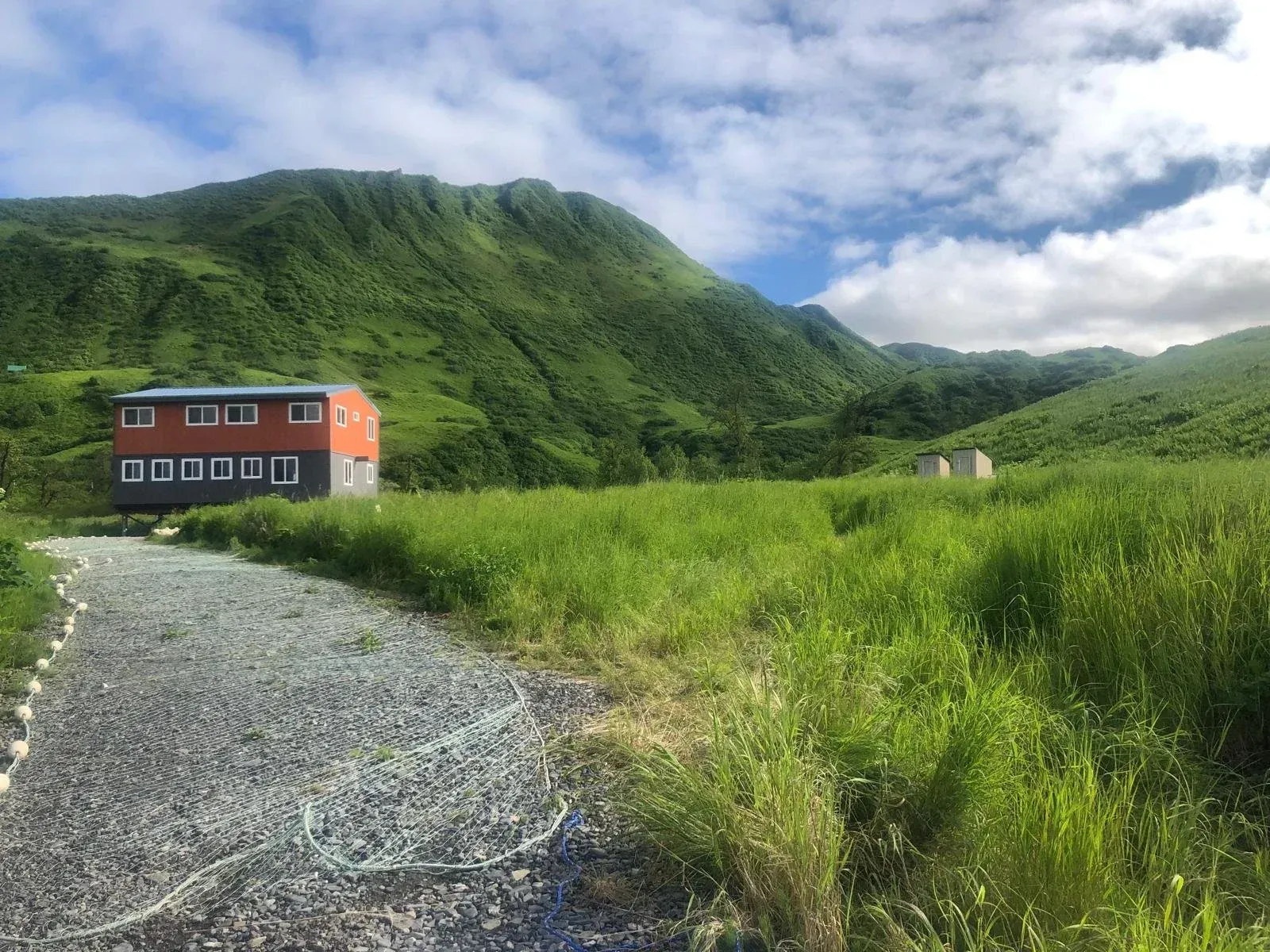 Two-story orange building with gray gravel path, grassy field, and green hills under cloudy sky.