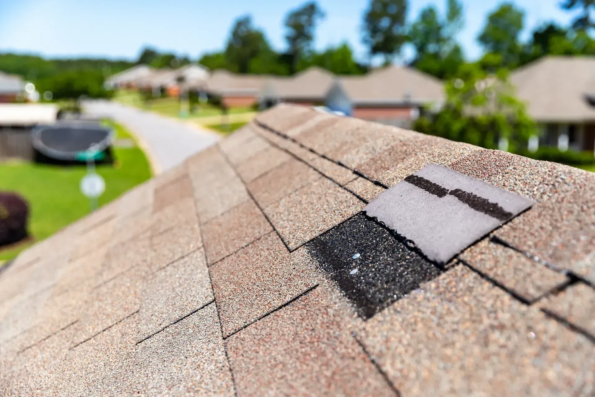 Close-up of a damaged asphalt shingle roof with a missing piece, overlooking a suburban street.