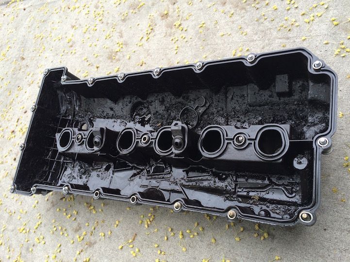 Black engine valve cover, oily residue inside, with round gasket openings and mounting bolts.