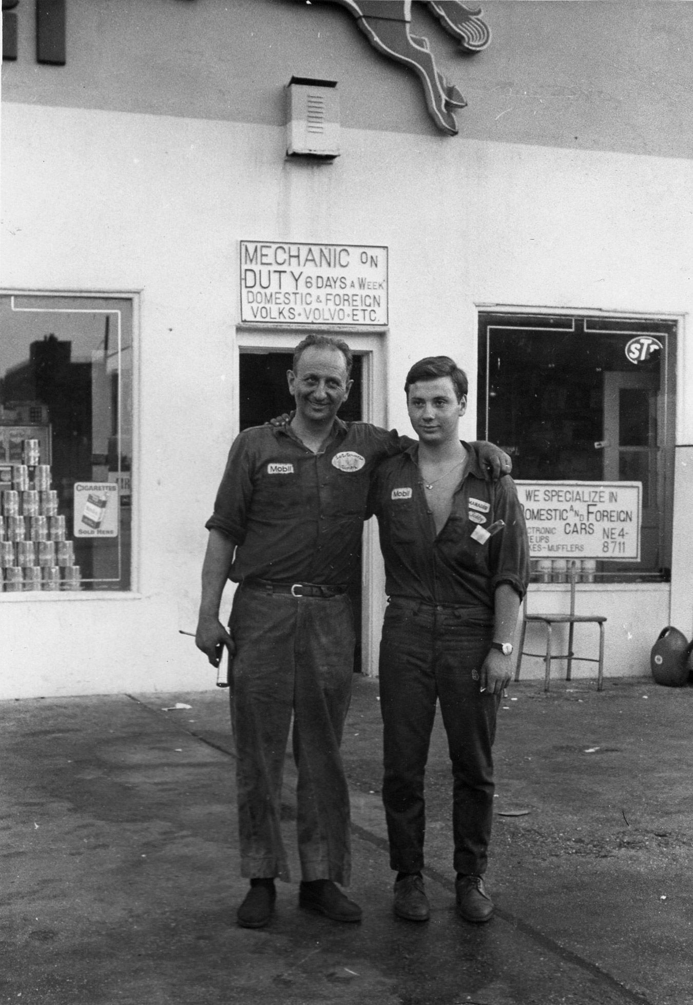 Two men in mechanic uniforms stand outside a gas station. One has arm around the other's shoulders. Sign says 