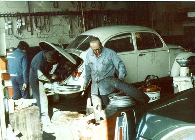 Mechanics working on a white VW Beetle in a garage. Two men with open hood, another man near the car.