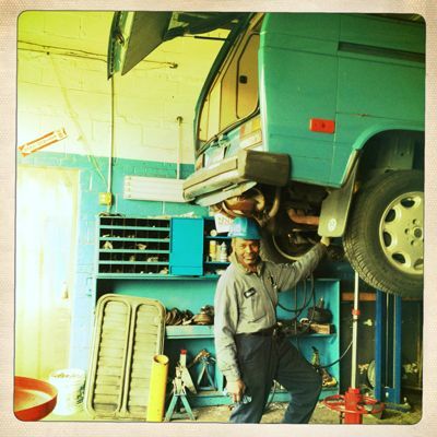 Mechanic in blue uniform under a green van, smiling, in a garage.
