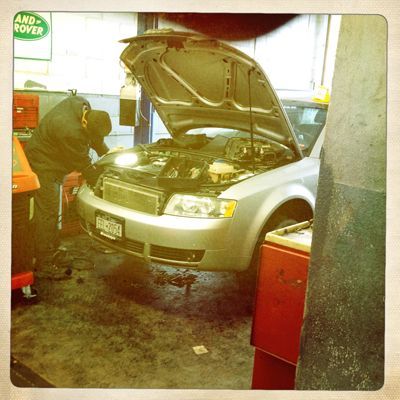 Two mechanics working on the open hood of a silver car in a garage.
