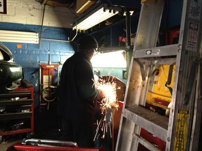 Mechanic grinding metal, creating sparks in a garage setting. Ladder and tools are nearby.